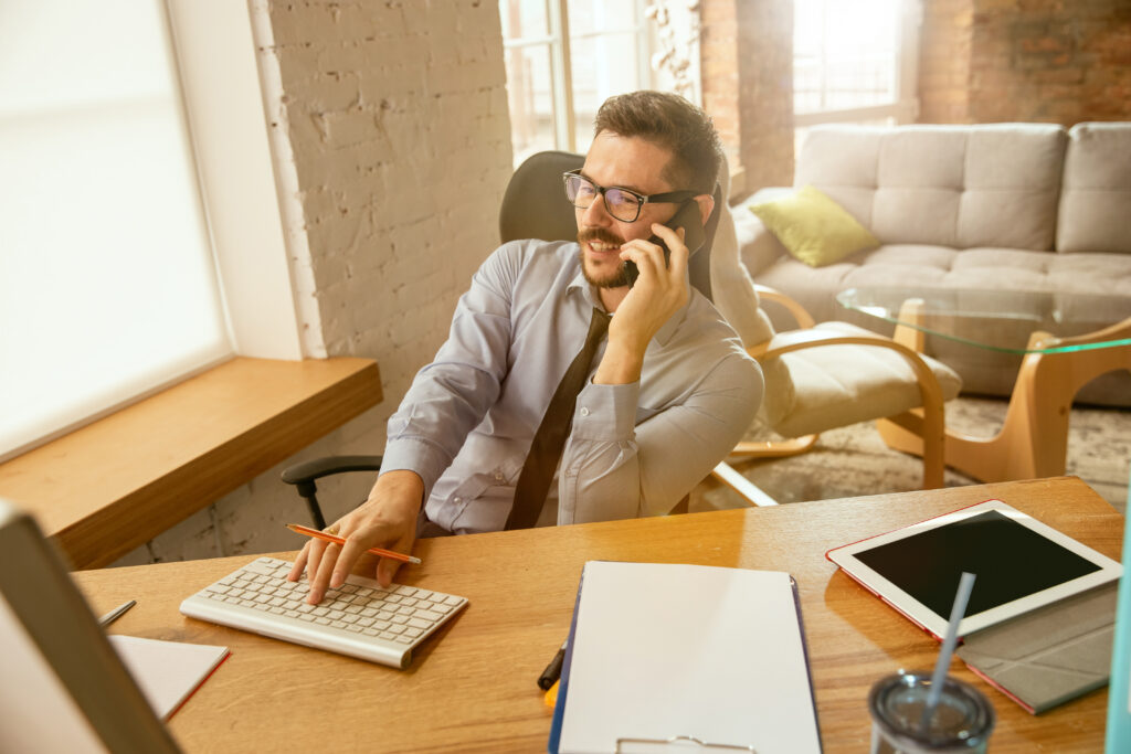 Mężczyzna w okularach rozmawia przez telefon komórkowy w jasnym, nowoczesnym biurze, obok niego klawiatura, notatnik i tablet.