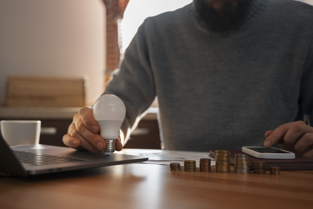 Mężczyzna trzymający żarówkę LED przy biurku z laptopem i stosami monet, symbolizując oszczędności energii i planowanie finansowe, dla strony firmy Direct4Energy.