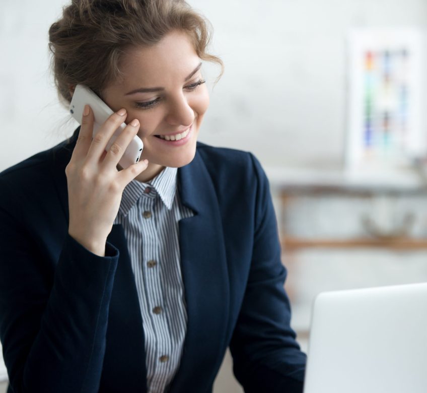 Portrait of beautiful happy smiling young architect woman talking on phone and working on laptop at home office desk. Attractive cheerful model using smartphone and computer, making call, taking order